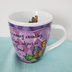 May include: Purple and green ceramic coffee mug with a diamond pattern. The mug has a white handle and features a cartoon illustration of stacks of coins and dollar bills with the text "It's a money counting, wishing, subject to  fund thing".