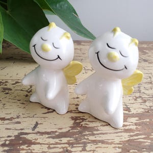 Op de afbeelding: Twee witte keramische beeldjes met lachende gezichten, gele accenten en kleine gele vleugels. De beeldjes hebben gesloten ogen en staan op een verweerd houten oppervlak. De beeldjes zijn decoratief.