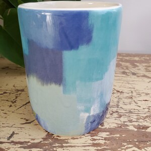 K&ouml;nnte beinhalten: Ein Keramikbecher mit handbemaltem Design. Der Becher zeigt eine Palette von Blau- und T&uuml;rkist&ouml;nen in einem Pinselstrichmuster. Der Innenraum ist wei&szlig;, und der Becher steht auf einer abgenutzten Holzoberfl&auml;che.
