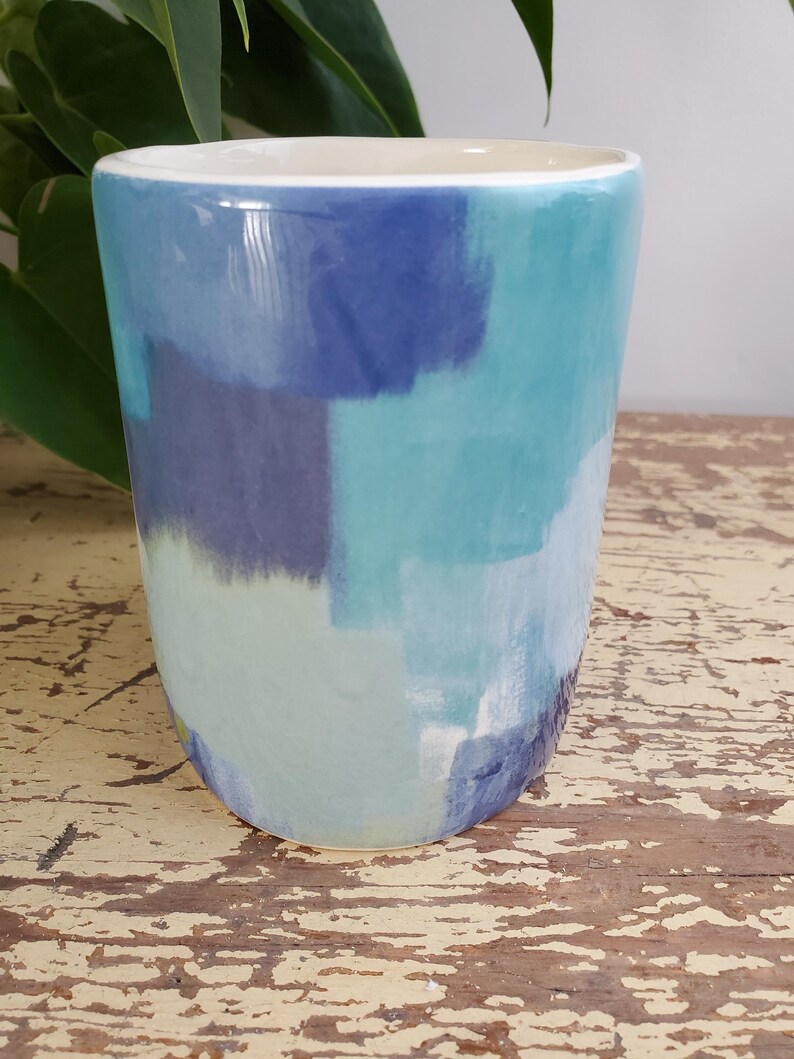 K&ouml;nnte beinhalten: Ein Keramikbecher mit handbemaltem Design. Der Becher zeigt eine Palette von Blau- und T&uuml;rkist&ouml;nen in einem Pinselstrichmuster. Der Innenraum ist wei&szlig;, und der Becher steht auf einer abgenutzten Holzoberfl&auml;che.
