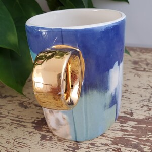 K&ouml;nnte beinhalten: Eine Keramiktasse mit goldfarbenem Griff. Die Tasse hat ein blau-wei&szlig;es Maldesign. Der Innenraum ist wei&szlig;. Die Tasse steht auf einer alten Holzoberfl&auml;che.