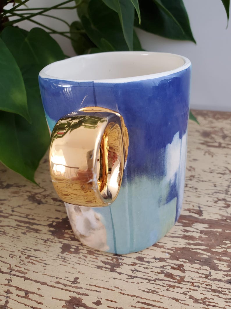 K&ouml;nnte beinhalten: Eine Keramiktasse mit goldfarbenem Griff. Die Tasse hat ein blau-wei&szlig;es Maldesign. Der Innenraum ist wei&szlig;. Die Tasse steht auf einer alten Holzoberfl&auml;che.