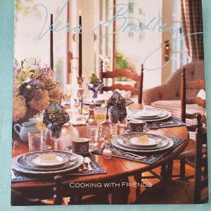 May include: A cookbook titled "Vera Bradley Cooking with Friends" featuring a photograph of a dining table set with blue and white patterned china, silverware, and glasses. The table is surrounded by chairs and a window with white curtains.