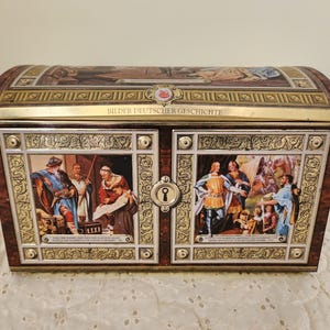 May include: A vintage decorative chest with a domed lid. The chest features illustrations of historical figures and the text "Bilder Deutscher Geschichte". The chest has a dark brown wood grain pattern with gold trim.