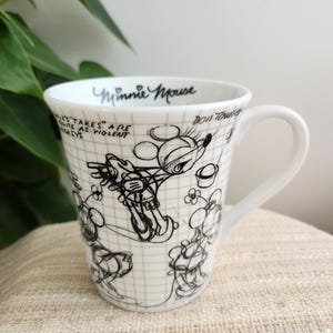 Op de afbeelding: Witte keramische mok met een zwart-witte schets van Minnie Mouse. De mok heeft een oor en is versierd met een rasterpatroon en tekst met de tekst "Minnie Mouse" en andere handgeschreven zinnen. Een klassiek Disney-verzamelobject.