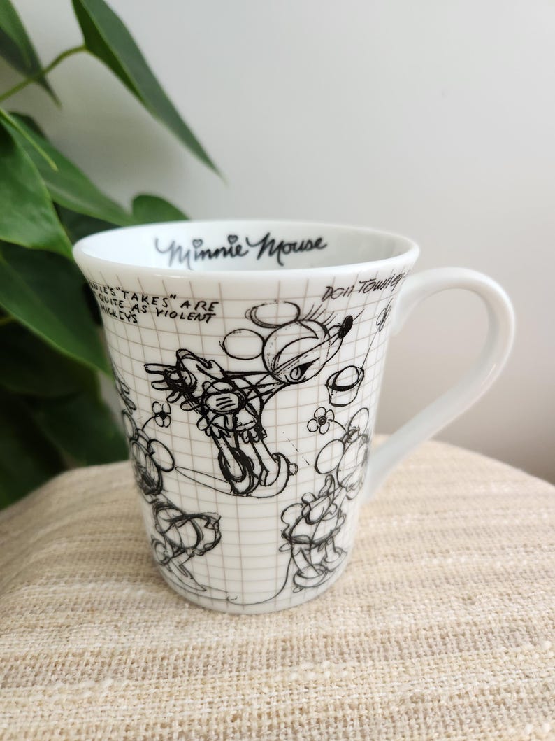 Op de afbeelding: Witte keramische mok met een zwart-witte schets van Minnie Mouse. De mok heeft een oor en is versierd met een rasterpatroon en tekst met de tekst "Minnie Mouse" en andere handgeschreven zinnen. Een klassiek Disney-verzamelobject.