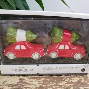 Op de afbeelding: Een set rode zout- en peperstrooiers in de vorm van een auto, elk met een groene kerstboom erop. De strooiers zitten in een doorzichtige plastic doos met de tekst "SALT & PEPPER SHAKER SET" en het merk "THRESHOLD".