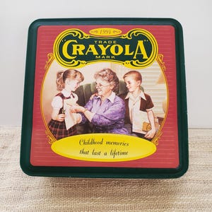 May include: A vintage green and red tin box with a picture of a grandmother and two children. The box has the words "Crayola Trade Mark" and "Childhood memories that last a lifetime" printed on it.