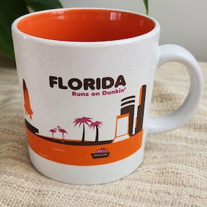 Könnte beinhalten: Ein weißer Keramikbecher mit leuchtend orangefarbenem Innenraum. Die Außenseite zeigt den Text FLORIDA Runs on Dunkin' in Braun und Pink, begleitet von einer grafischen Darstellung eines Space Shuttles, rosa Flamingos, Palmen und einem Boot.