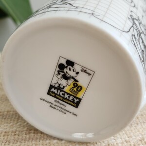 Op de afbeelding: Witte keramische mok met een zwart-wit schets van Mickey Mouse en een rasterpatroon. De mok heeft een Disney-logo met de tekst "90 Years of Magic" en "Mickey The True Original". Vaatwasser- en magnetronbestendig.