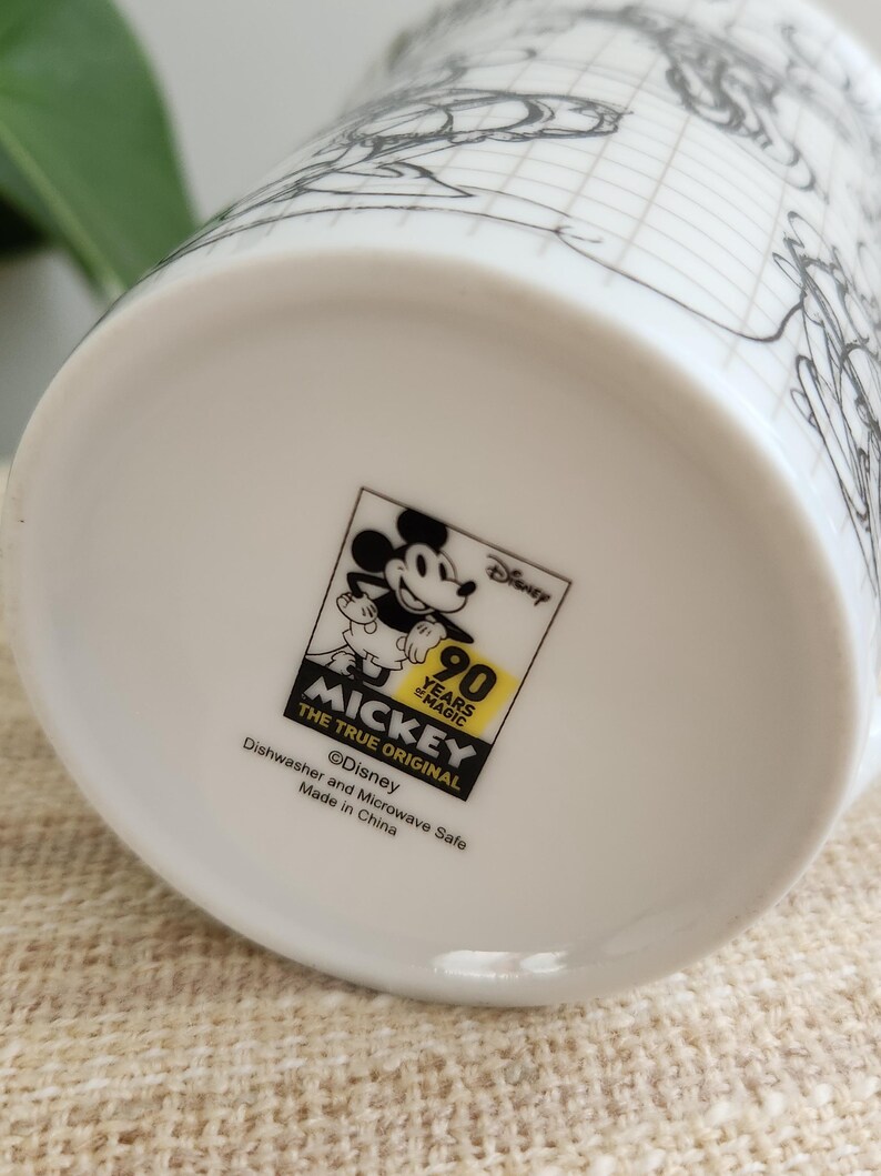 Op de afbeelding: Witte keramische mok met een zwart-wit schets van Mickey Mouse en een rasterpatroon. De mok heeft een Disney-logo met de tekst "90 Years of Magic" en "Mickey The True Original". Vaatwasser- en magnetronbestendig.