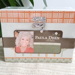 May include: A recipe card box with a white and orange plaid pattern on the lid. The box features a silver clasp and a label with the text "Paula Deen Kitchen" and "Recipe Card Box". The box is wrapped in clear plastic.