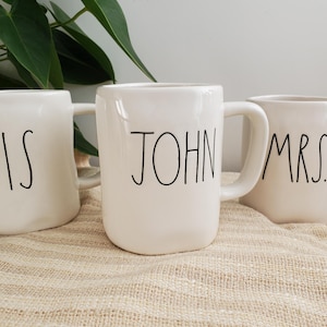 Puede incluir: Tres tazas de cerámica blanca con texto negro. Una taza dice "HIS", otra "JOHN" y la tercera "MRS.". Las tazas tienen un diseño sencillo y moderno con un asa curvada.