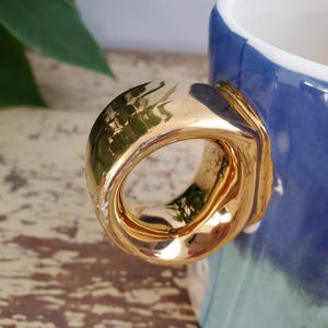 K&ouml;nnte beinhalten: Nahaufnahme einer Keramiktasse mit einem goldfarbenen Henkel. Die Tasse hat eine blau-wei&szlig;e glasierte Au&szlig;enseite. Der Henkel hat eine dicke, abgerundete Form mit gl&auml;nzendem Finish. Das Design der Tasse ist schlicht und elegant.