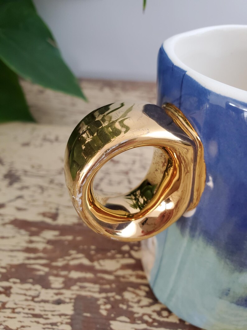 K&ouml;nnte beinhalten: Nahaufnahme einer Keramiktasse mit einem goldfarbenen Henkel. Die Tasse hat eine blau-wei&szlig;e glasierte Au&szlig;enseite. Der Henkel hat eine dicke, abgerundete Form mit gl&auml;nzendem Finish. Das Design der Tasse ist schlicht und elegant.