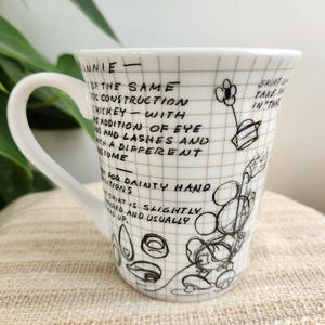 Op de afbeelding: Witte keramische mok met een handvat, met een zwart-witte schets van Minnie Mouse en tekst. De mok heeft een rasterpatroon en handgeschreven notities over het ontwerp van het personage. De mok staat op een beige oppervlak.