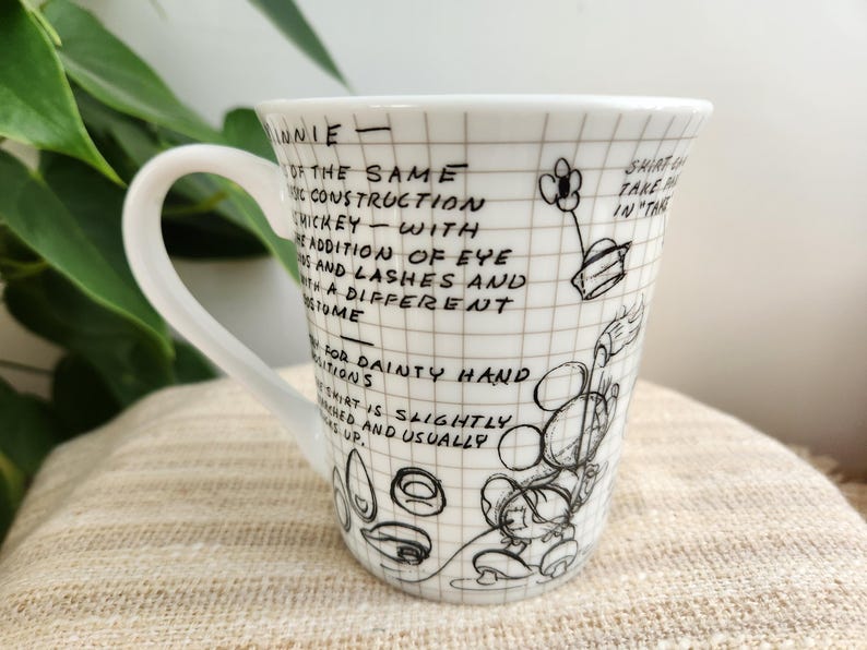 Op de afbeelding: Witte keramische mok met een handvat, met een zwart-witte schets van Minnie Mouse en tekst. De mok heeft een rasterpatroon en handgeschreven notities over het ontwerp van het personage. De mok staat op een beige oppervlak.