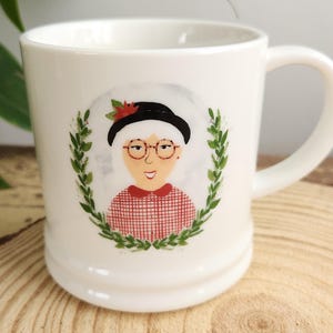 Op de afbeelding: Witte keramische mok met een illustratie van een vrouw met een bril, een zwarte hoed en een rood-wit geruit overhemd, omringd door een groene krans. De mok heeft een gebogen handvat en een gladde, glanzende afwerking.