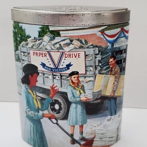 May include: A vintage tin container with a colorful illustration of two Girl Scouts collecting newspapers for a "Paper Drive for Victory" campaign. The illustration features a truck filled with newspapers, a post office, and a red, white, and blue banner.