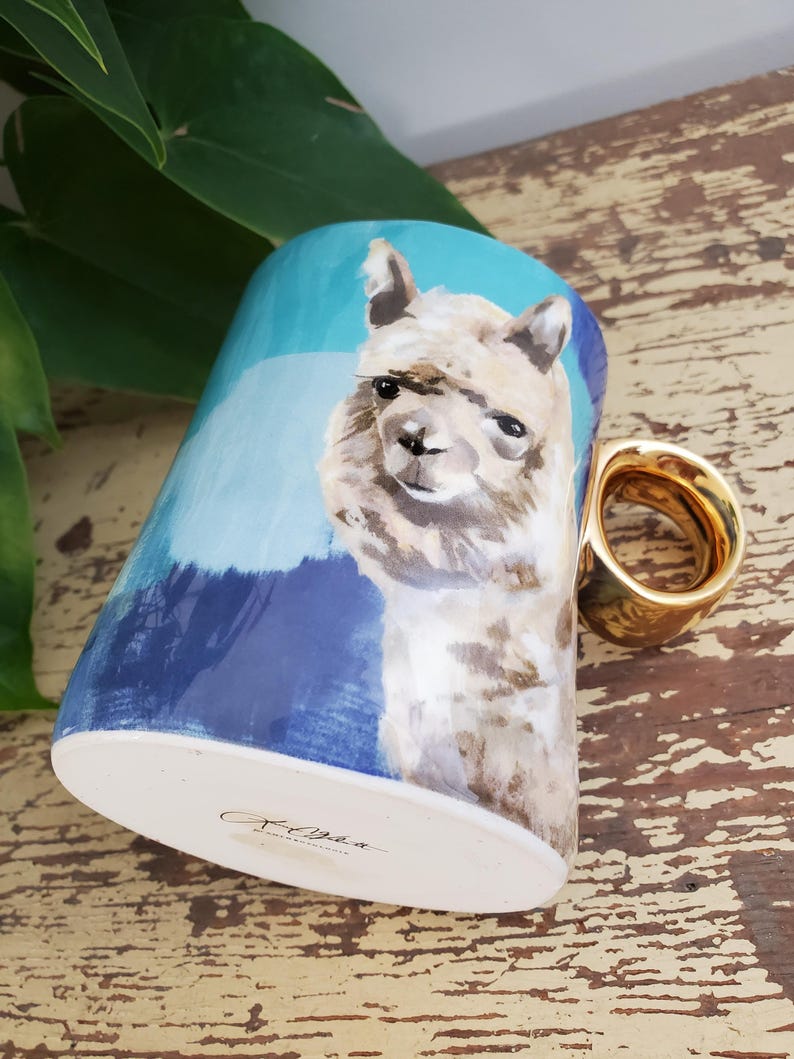 K&ouml;nnte beinhalten: Keramikbecher mit handgemaltem Alpaka-Design und goldenem Henkel. Der Becher hat einen blau-wei&szlig;en Hintergrund. Der Boden des Bechers hat eine Signatur mit schwarzer Tinte. Der Becher steht auf einer Holzoberfl&auml;che.