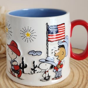 May include: A white ceramic mug with a bright red handle and a light blue interior. The exterior features a raised, embossed design of Peanuts characters including Snoopy and Charlie Brown wearing cowboy hats, a desert landscape, and an American flag.