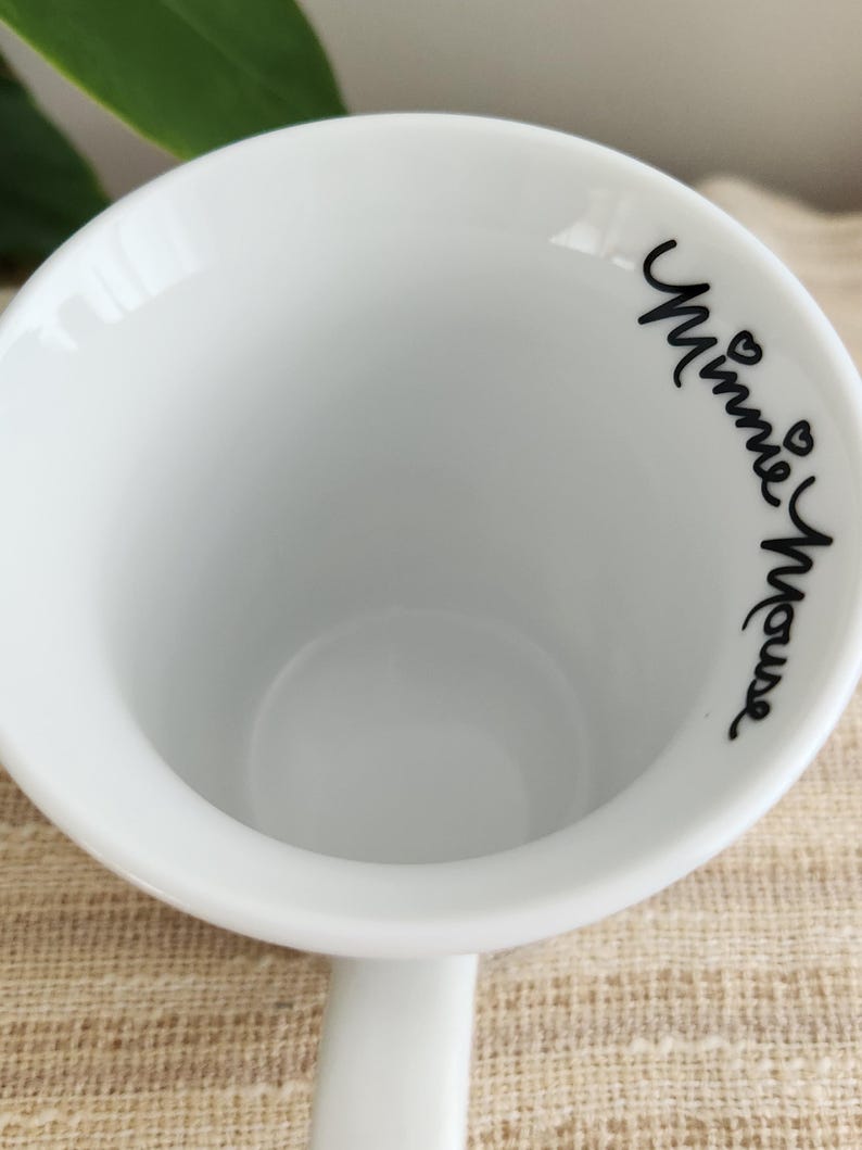 Op de afbeelding: Witte keramische mok met de woorden "Minnie Mouse" in zwart script op de rand. De mok heeft een eenvoudig, klassiek ontwerp met een afgeronde body en een handvat. De mok staat op een beige oppervlak.