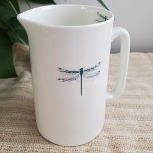 May include: White ceramic pitcher with a handle and a spout. The pitcher is decorated with a teal dragonfly design on the front and inside the rim. The pitcher is sitting on a beige textured surface.