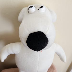May include: A white plush dog toy with a large, rounded black nose and plastic eyes that are positioned looking upward. The character has a smooth, soft texture, floppy ears, and a simple, rounded body shape with two arms and two feet visible.