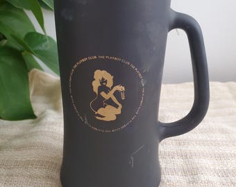 Peut inclure: Un grand mug en céramique noire avec une anse. Le mug présente un dessin doré d'une femme et le texte "THE PLAYBOY CLUB" dans un cercle. Le mug est posé sur une surface beige.