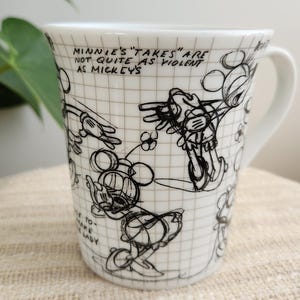 Op de afbeelding: Witte keramische mok met zwarte schetsen van Mickey en Minnie Mouse. De mok heeft een oor en is versierd met cartoontekeningen en de tekst "MINNIE'S 'TAKES' ARE NOT QUITE AS VIOLENT AS MICKEY'S".