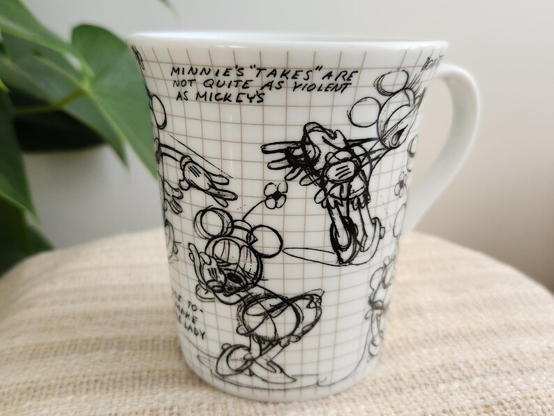 Op de afbeelding: Witte keramische mok met zwarte schetsen van Mickey en Minnie Mouse. De mok heeft een oor en is versierd met cartoontekeningen en de tekst "MINNIE'S 'TAKES' ARE NOT QUITE AS VIOLENT AS MICKEY'S".