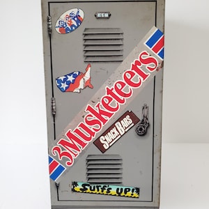 Puede incluir: Un casillero de metal gris con varias pegatinas, incluyendo una pegatina de la barra de chocolate 3 Mosqueteros con el texto "Snack Bars", una pegatina con el texto "Surfs Up!" y una pegatina con una representación de la bandera de los Estados Unidos.