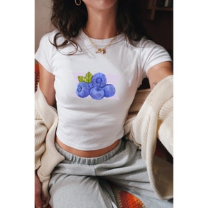 May include: White crop top with a watercolor illustration of blueberries on a light purple background.