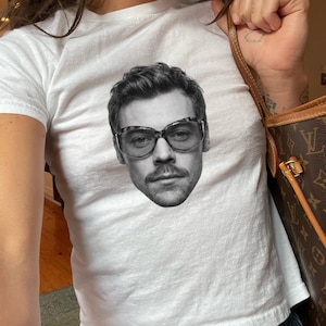 May include: A white short-sleeved t-shirt featuring a centered black and white photographic print of a person with a mustache wearing thick-rimmed glasses. The graphic is positioned on the chest area of the fitted cotton crew neck garment.
