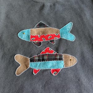 May include: Dark gray sweatshirt with two appliqued fish. The fish have brown, red, and plaid patterned bodies with blue tail fins. The fish are outlined with white stitching and have gold-colored accents.