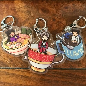 May include: Three clear acrylic keychains featuring anime characters. The first keychain shows a girl with purple hair in a bowl of ramen. The second keychain shows a boy with brown hair in a red teacup. The third keychain shows a boy with brown hair in a blue coffee mug with the text "#2 LAWYER".
