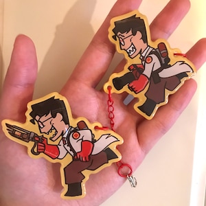 May include: Two cartoon keyrings depicting a character in a white lab coat, red gloves, and brown trousers, holding a weapon. The keyrings are linked by a red chain and have a yellow background. The character has black hair and glasses.