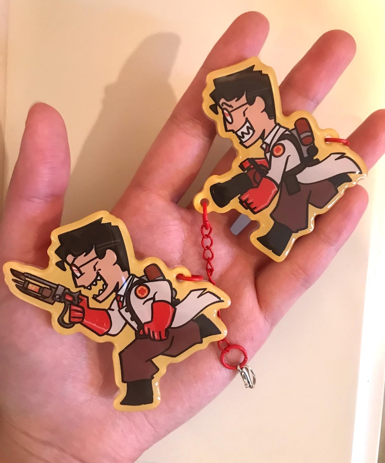 May include: Two cartoon keyrings depicting a character in a white lab coat, red gloves, and brown trousers, holding a weapon. The keyrings are linked by a red chain and have a yellow background. The character has black hair and glasses.