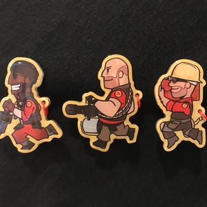 May include: Three cartoon character pins. Each pin depicts a different character in a running pose, with a red and brown colour scheme. The pins have a yellow border and a small red string attached.