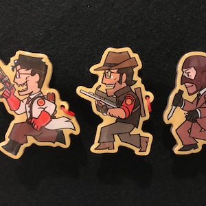 May include: Three cartoon character keyrings. The first is a doctor in a white coat and red gloves holding a weapon. The second is a man in a brown hat holding a rifle. The third is a man in a suit holding a knife.