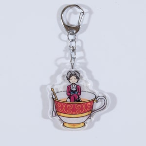 May include: A clear acrylic keychain featuring a cartoon character sitting in a red and gold teacup. The character has gray hair and is wearing a red jacket and blue pants.
