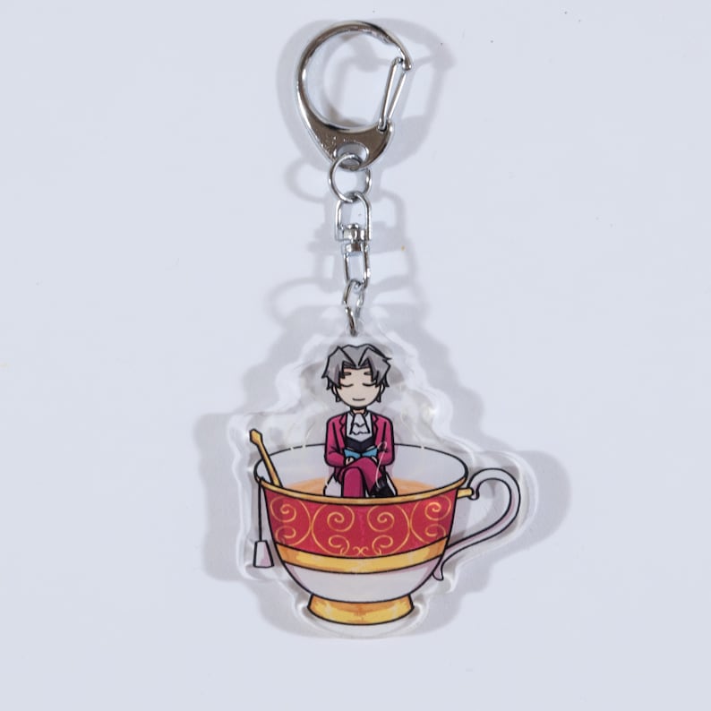 May include: A clear acrylic keychain featuring a cartoon character sitting in a red and gold teacup. The character has gray hair and is wearing a red jacket and blue pants.