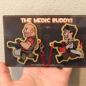 May include: A decorative plaque featuring two cartoon characters from the video game Team Fortress 2. The characters are the Heavy and the Medic, each holding a weapon. The plaque has the text "THE MEDIC BUDDY!" and is connected by a red chain.