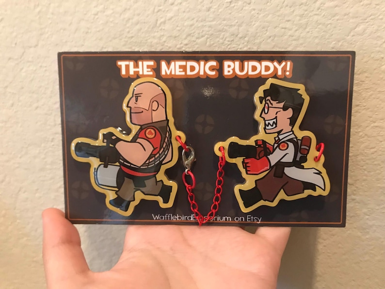 May include: A decorative plaque featuring two cartoon characters from the video game Team Fortress 2. The characters are the Heavy and the Medic, each holding a weapon. The plaque has the text "THE MEDIC BUDDY!" and is connected by a red chain.