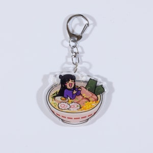 May include: A clear acrylic keychain featuring a cartoon illustration of a girl sitting in a bowl of ramen. The girl is wearing a purple shirt and has black hair. The ramen bowl is red and white with a variety of toppings, including a boiled egg, green onions, and narutomaki.
