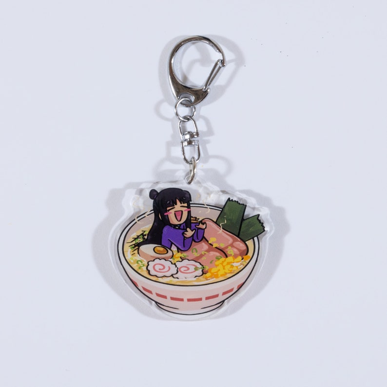 May include: A clear acrylic keychain featuring a cartoon illustration of a girl sitting in a bowl of ramen. The girl is wearing a purple shirt and has black hair. The ramen bowl is red and white with a variety of toppings, including a boiled egg, green onions, and narutomaki.