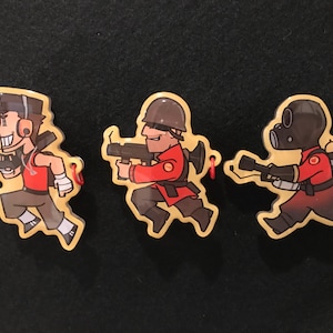 May include: Three cartoon keyrings featuring characters from Team Fortress 2. The characters are depicted running and holding weapons. They are wearing red, brown, and black outfits. Each keyring has a red string.