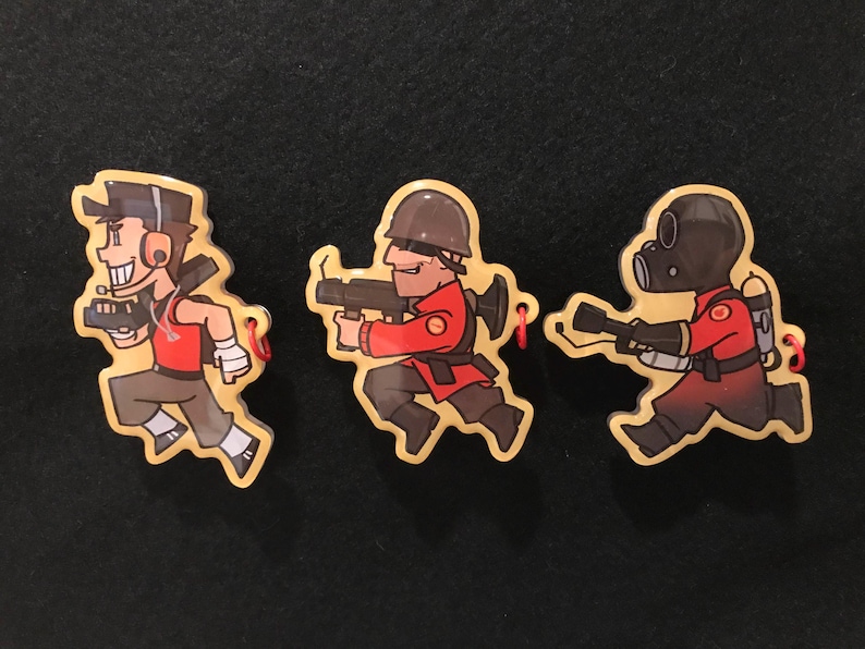 May include: Three cartoon keyrings featuring characters from Team Fortress 2. The characters are depicted running and holding weapons. They are wearing red, brown, and black outfits. Each keyring has a red string.