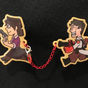 May include: Two cartoon character keyrings linked by a red chain. One depicts a woman in a purple shirt and black skirt holding a gun. The other is a man in a white coat and red gloves, also with a gun.