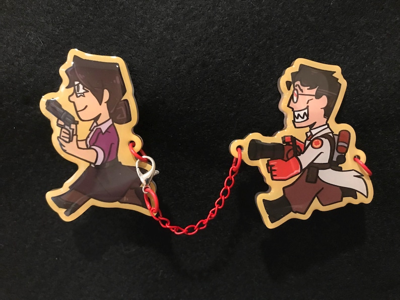 May include: Two cartoon character keyrings linked by a red chain. One depicts a woman in a purple shirt and black skirt holding a gun. The other is a man in a white coat and red gloves, also with a gun.